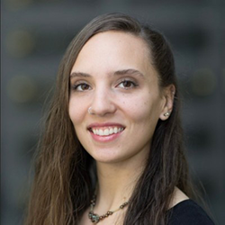 A woman smiling at the camera with brown hair, bown eyes a necklace and a black shirt.