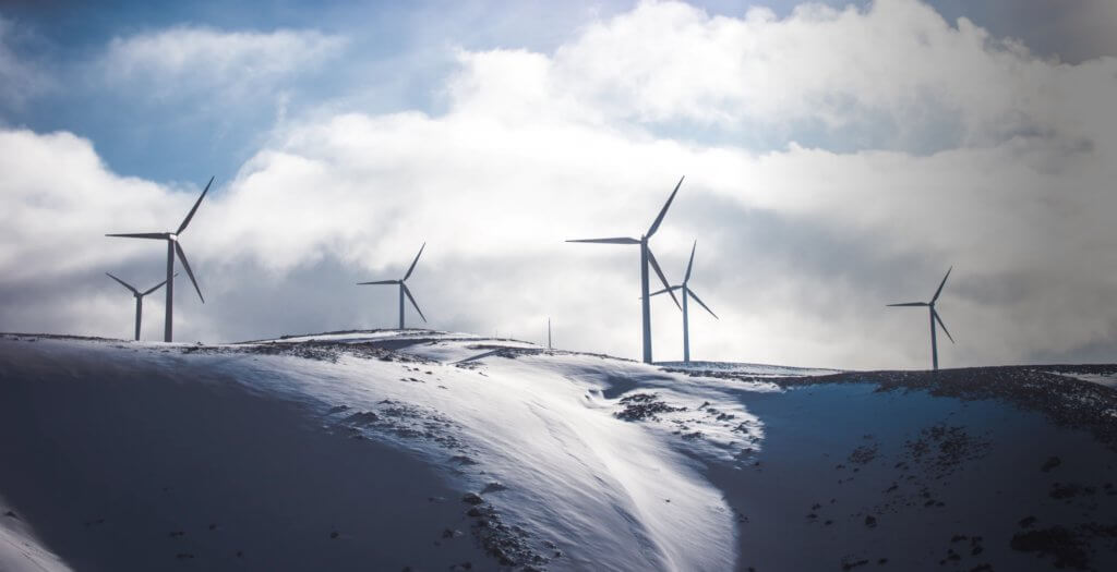 A wide-angle landscape image of wind turbines on a snowy mountaintop against a cloudy sky.