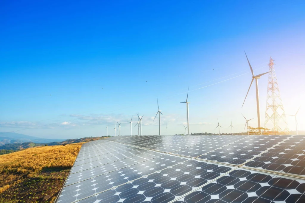 A row of solar panels in front of a row of wind turbines and transmission towers on a clear day.