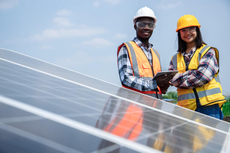 two adults wearing safety vests and hard hats smiling behind some solar panels