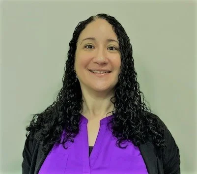 A headshot of a middle-aged woman with curly dark hair in a purple business casual shirt.