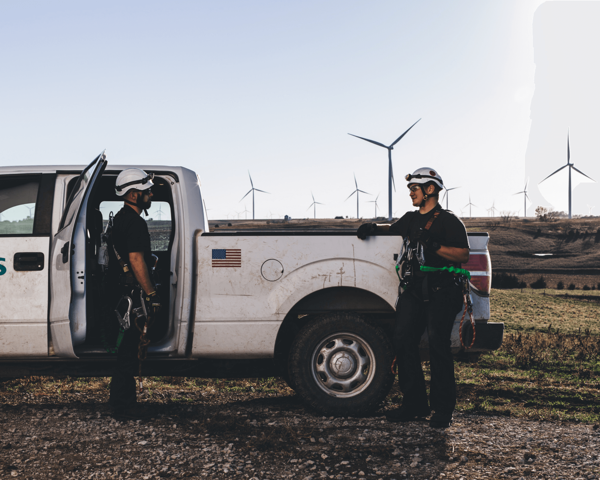 Two workers standing in front of a white pick-up truck with wind turbines in the background.