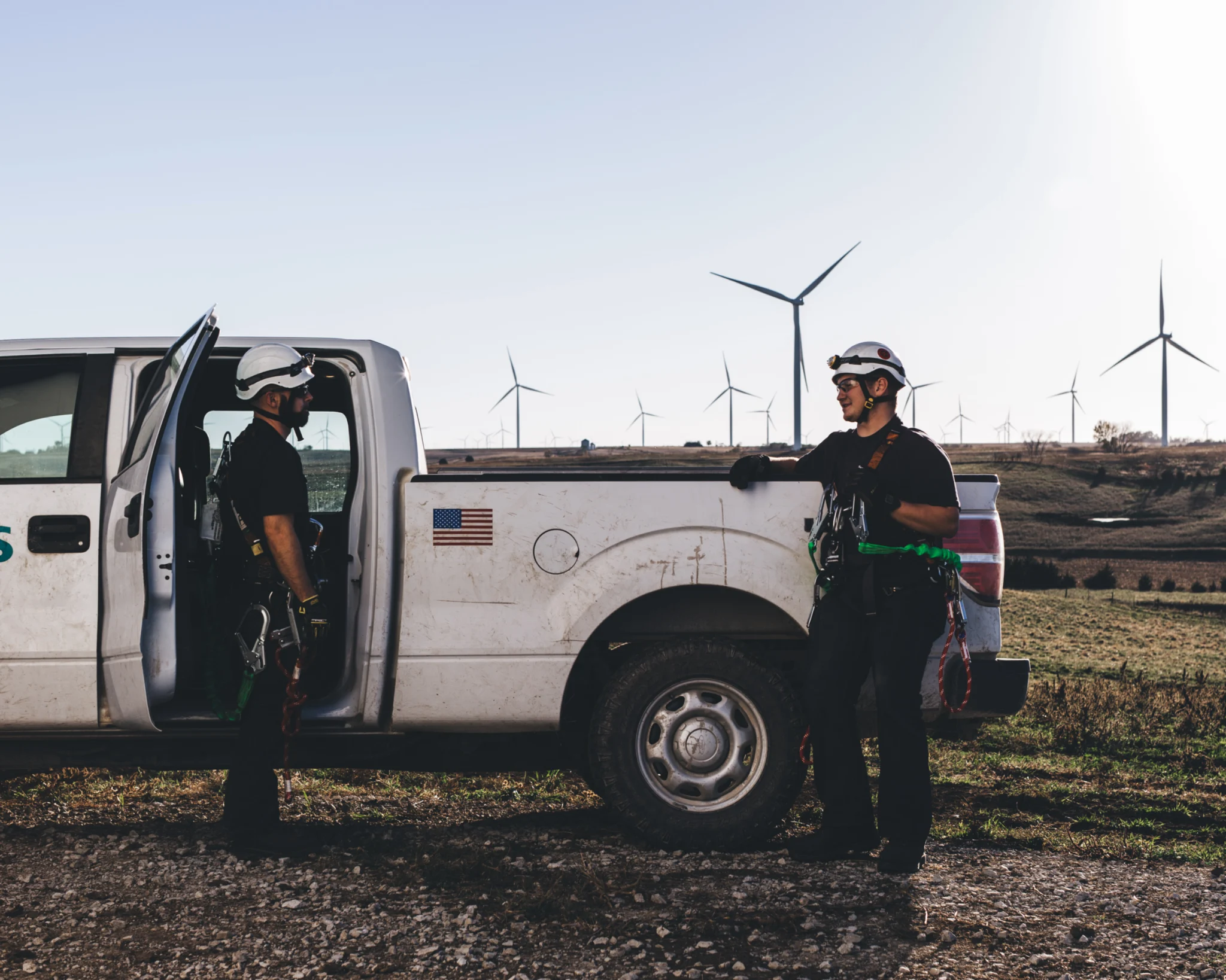 Two workers standing in front of a white pick-up truck with wind turbines in the background.