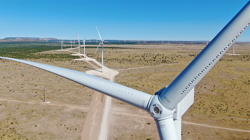 A photo of wind turbine blades in the foreground, with a row of turbines in the background along desert terrain.
