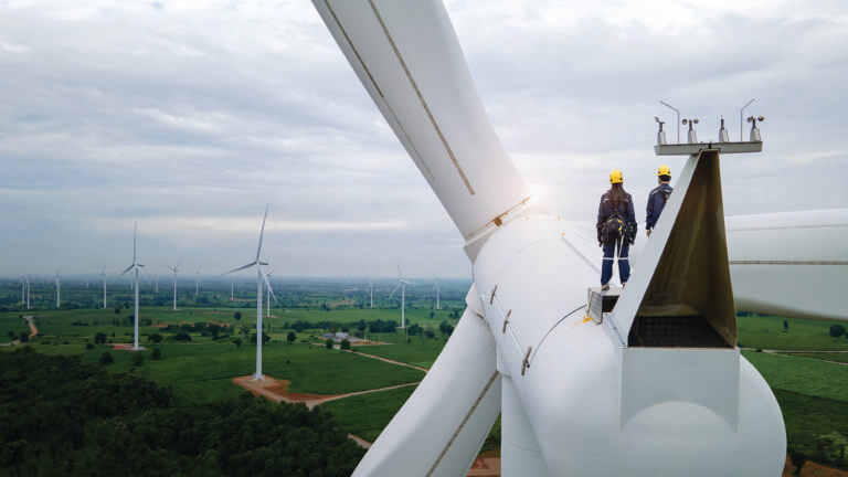 Two wind turbine technicians stand on top of a wind turbine, looking out on a field.