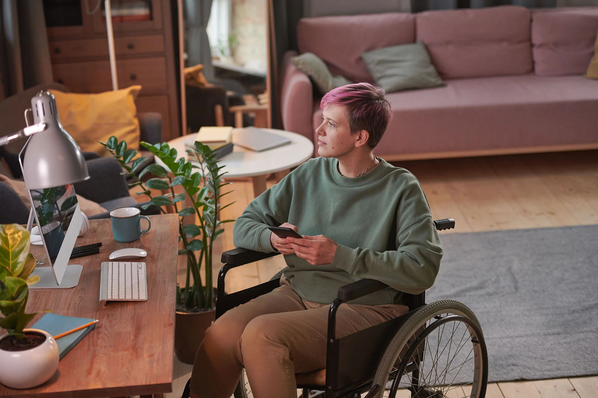 A young person with short pink hair sitting in wheelchair in front of her workplace and using her mobile phone