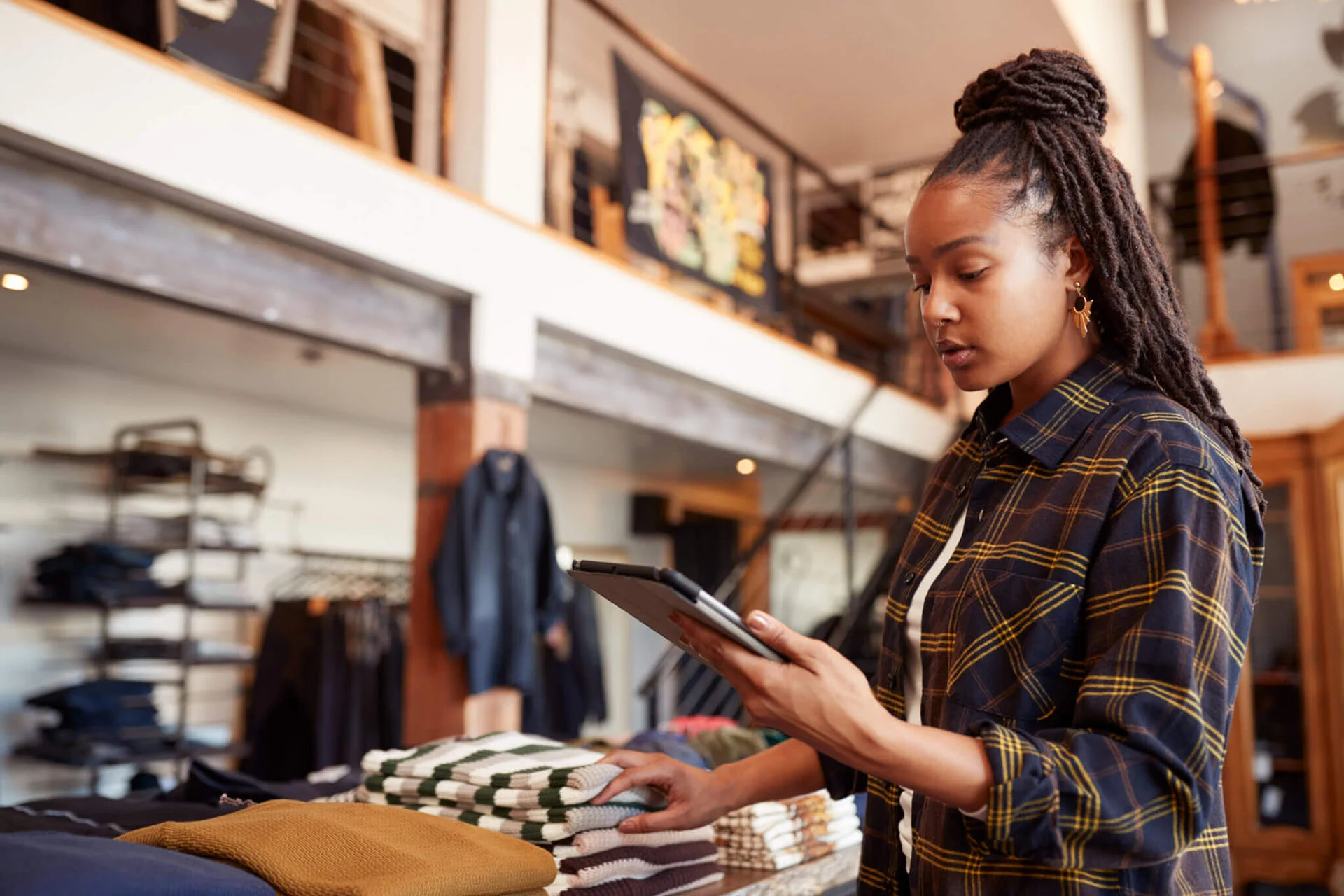 A young black woman with braids standing in front of a clothing store display looking at a tablet.