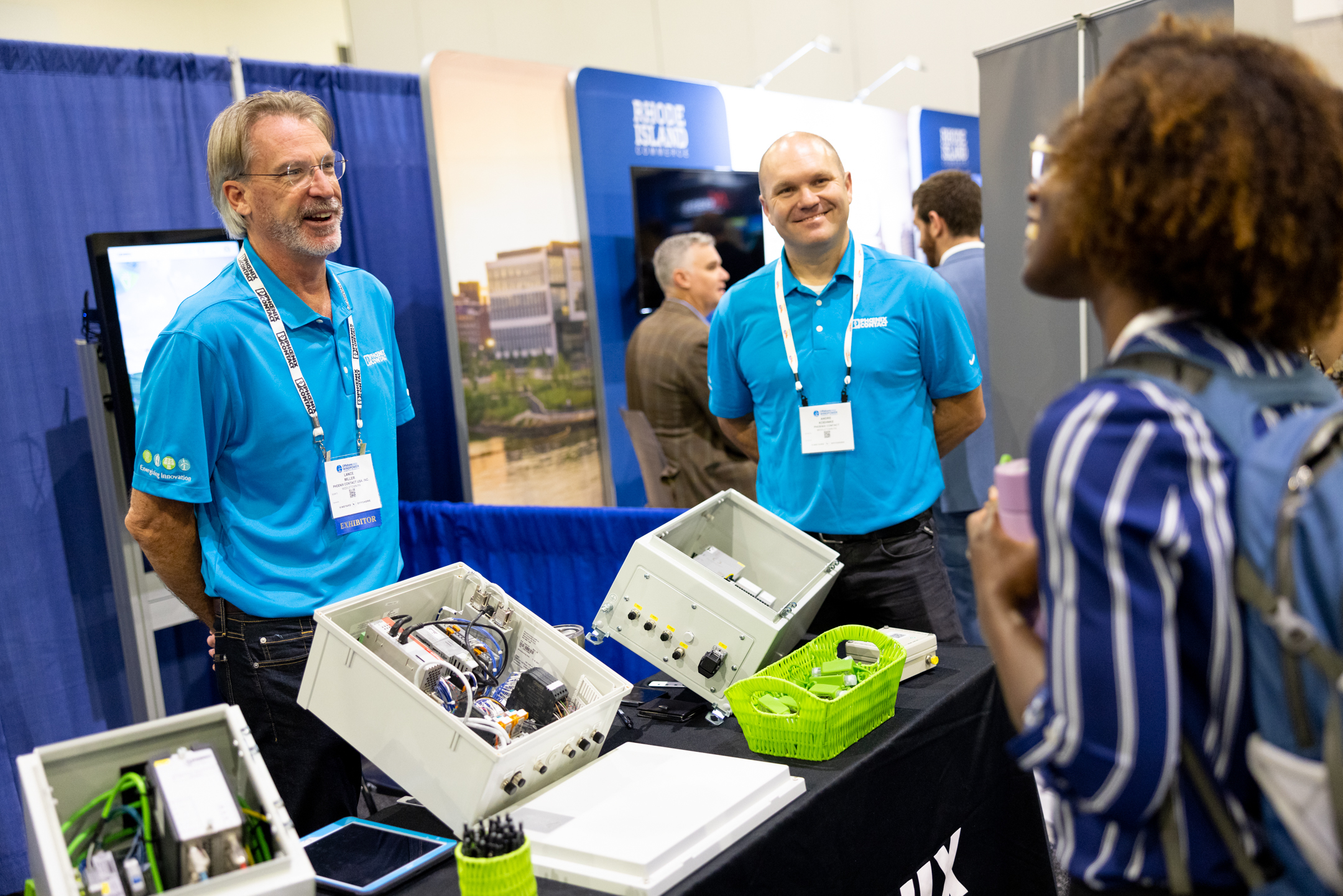 Two men wearing bright blue shirts on the exhibition floor at ACP's Offshore Windpower conference showing their product to a conference attendee to promote the Guess Watt challenge.