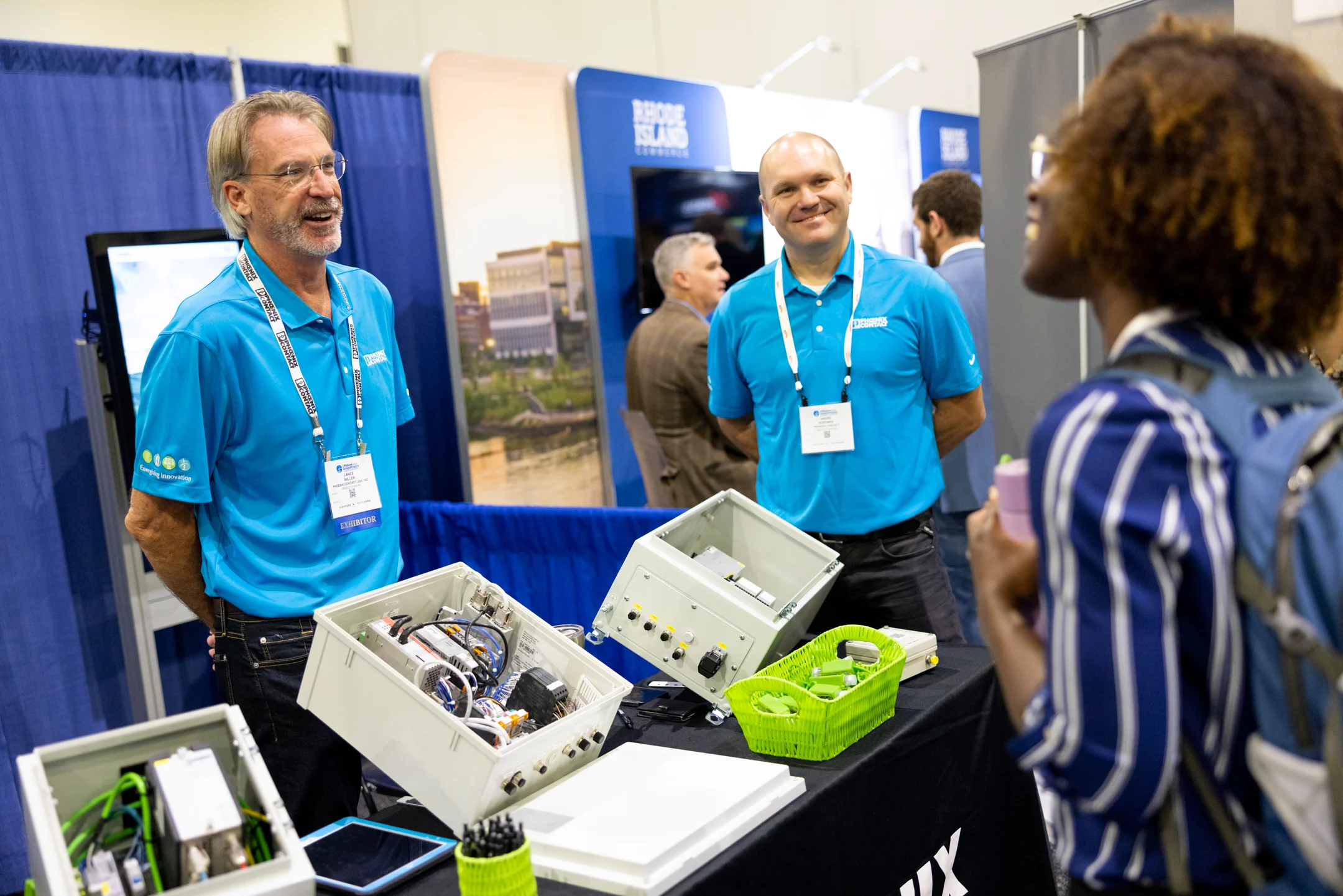 Two men wearing bright blue shirts on the exhibition floor at ACP's Offshore Windpower conference showing their product to a conference attendee to promote the Guess Watt challenge.