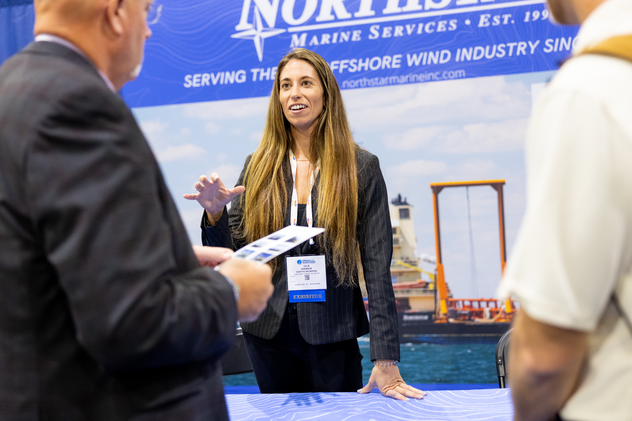 A woman exhibiting at ACP's Offshore Windpower conference discussing her organization with two attendees, wearing a lanyard and badge, two sponsorship opportunities.