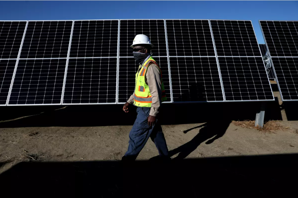 A man walking in front of a solar panel wearing a safety helmet and vest, originally published in an LA Times article referencing ACP California.