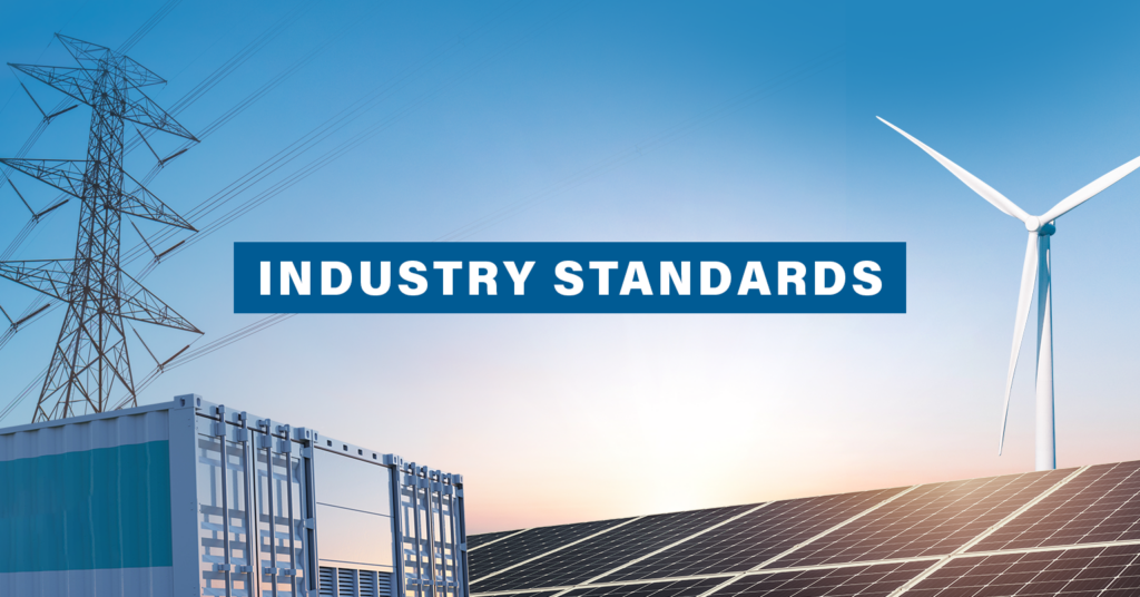 A graphic with the words "Industry Standards" in bold text hovering in the sky and in the background you can see solar panels, a wind turbine, an energy storage facility, and a transmission tower