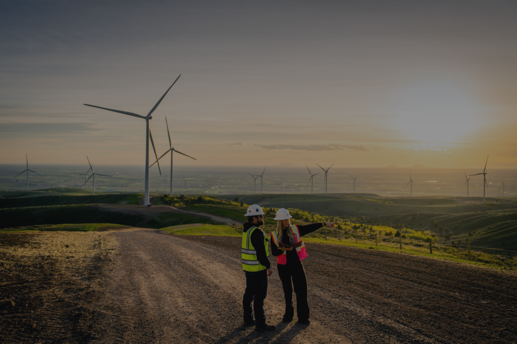 two people in safety gear standing together on a dirt path, one of them is pointing to the distance and the other looks in the direction of where they're pointing, a wind farm can be seen in the distant background