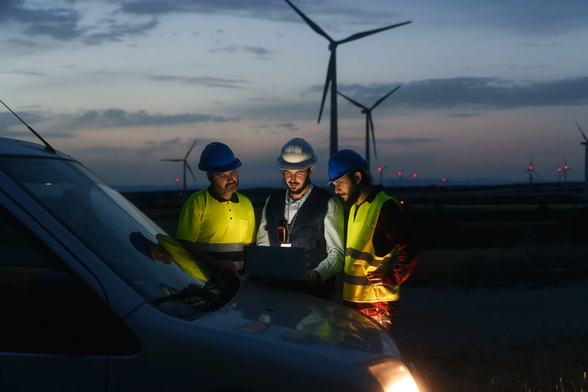 three workers wearing safety gear huddle around a computer on the hood of a car in the evening, a wind farm is visible in the background