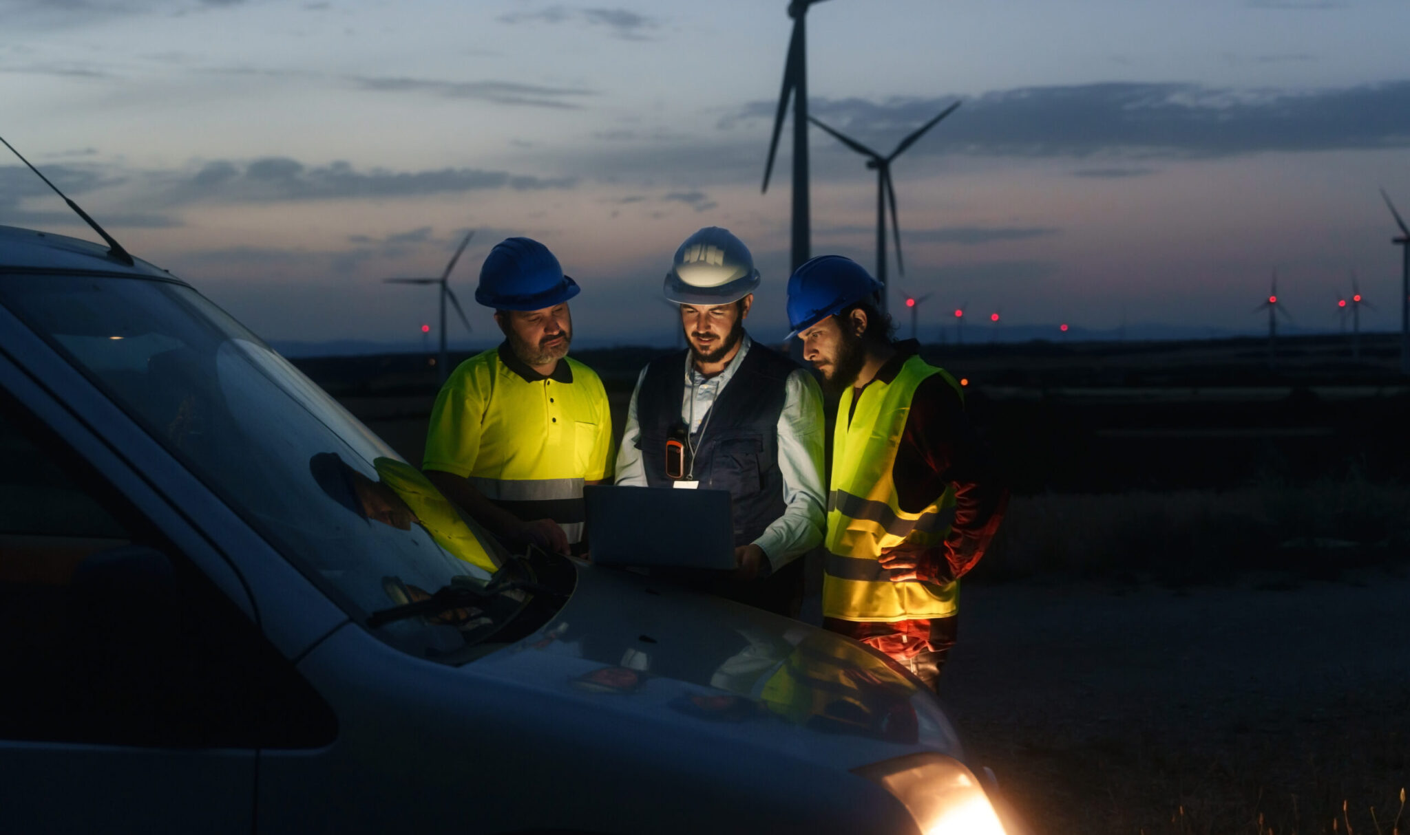 three workers wearing safety gear huddle around a computer on the hood of a car in the evening, a wind farm is visible in the background