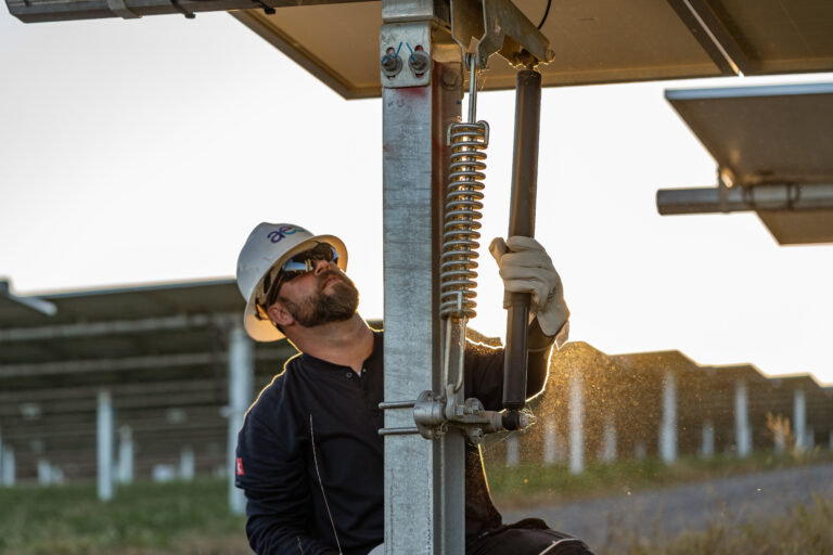 a worker wearing a hard hat, protective glasses and thick work gloves works on the spring below a solar panel