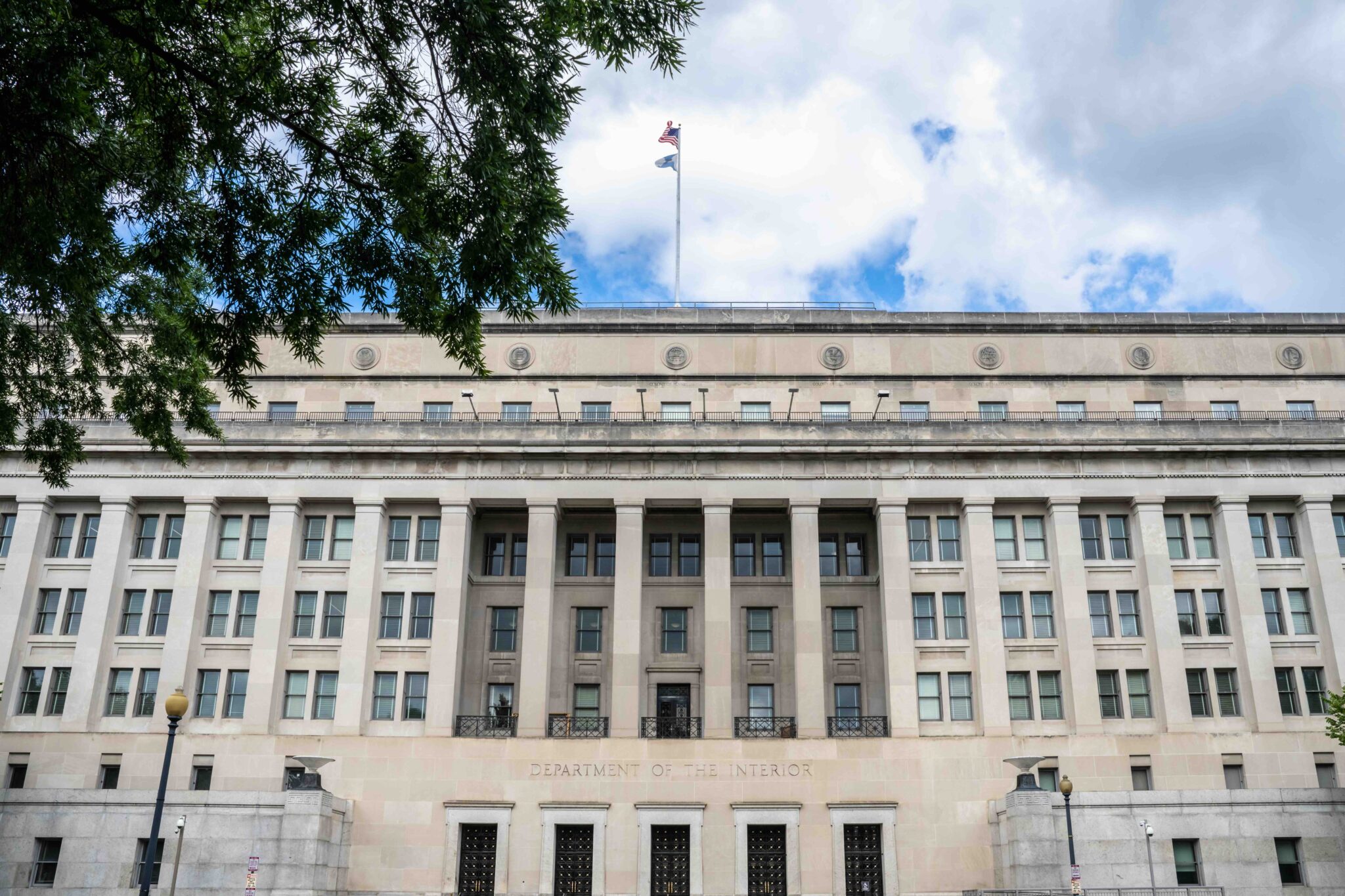 Front view of the U.S. Department of the Interior headquarters in Washington, D.C., framed by tree branches under a partly cloudy sky. The neoclassical building features tall columns and American and departmental flags flying from the rooftop.